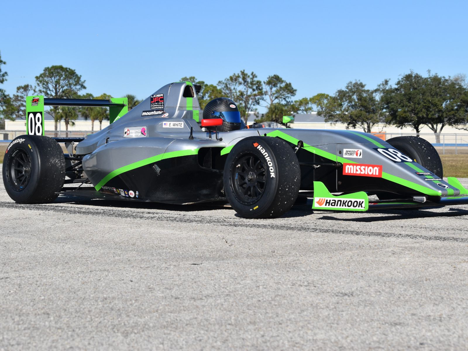 Silver and green open-wheel formula race car number 08 parked on a racetrack under clear sky