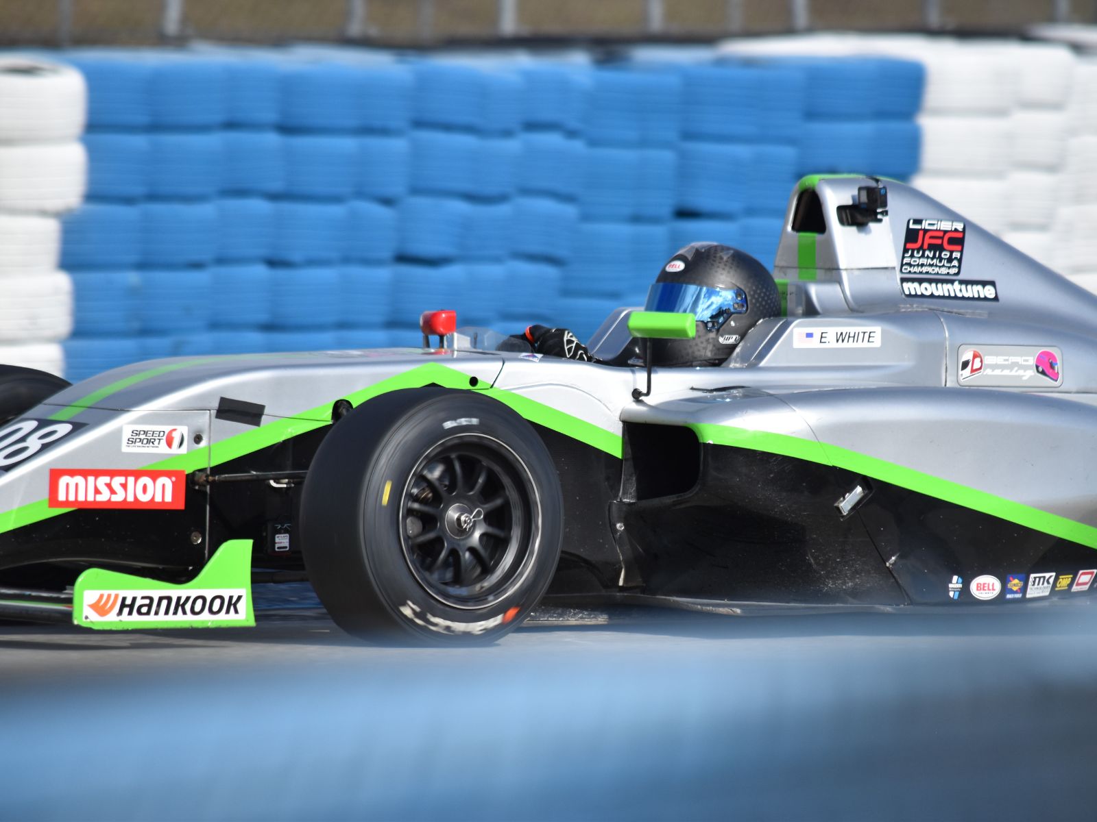 Open-wheel formula race car in silver and green livery driving past blue tire barriers on a race circuit